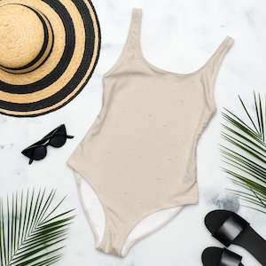 Peut inclure: Un maillot de bain une pi&egrave;ce beige avec un motif topographique subtil, accompagn&eacute; d'un chapeau de paille &agrave; rayures noires, de lunettes de soleil &oelig;il-de-chat noires et de sandales noires, dispos&eacute;s avec des palmes vertes sur un marbre blanc.