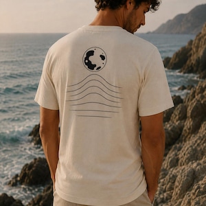 K&ouml;nnte beinhalten: Ein cremefarbenes Kurzarm-T-Shirt mit einer schwarzen Grafik auf der R&uuml;ckseite. Das Design besteht aus einer kreisf&ouml;rmigen Mondform &uuml;ber einer Reihe von f&uuml;nf horizontalen, wellenf&ouml;rmigen Linien, die sich in der Mitte nach oben w&ouml;lben.