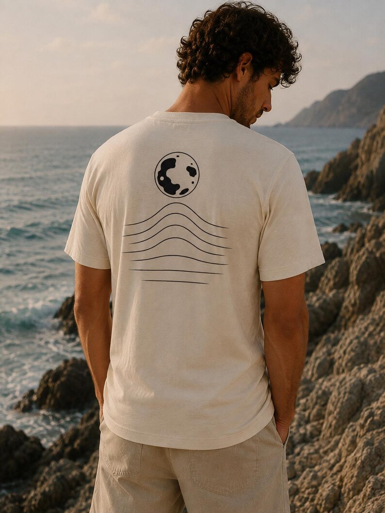K&ouml;nnte beinhalten: Ein cremefarbenes Kurzarm-T-Shirt mit einer schwarzen Grafik auf der R&uuml;ckseite. Das Design besteht aus einer kreisf&ouml;rmigen Mondform &uuml;ber einer Reihe von f&uuml;nf horizontalen, wellenf&ouml;rmigen Linien, die sich in der Mitte nach oben w&ouml;lben.