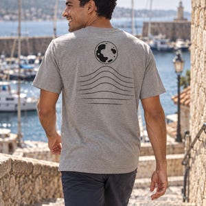 K&ouml;nnte beinhalten: Ein kurz&auml;rmeliges T-Shirt in Grau meliert mit einer schwarzen Grafik auf der R&uuml;ckseite. Das Design besteht aus einer kreisf&ouml;rmigen Mondform &uuml;ber einer Reihe von sechs horizontalen, wellenf&ouml;rmigen Linien, die nach unten hin breiter werden.