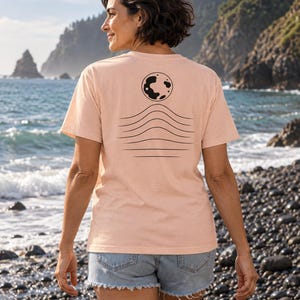 K&ouml;nnte beinhalten: Ein hellpfirsichfarbenes Kurzarm-T-Shirt mit einer schwarzen Grafik auf der R&uuml;ckseite. Das Design besteht aus einer kreisf&ouml;rmigen Mondform &uuml;ber einer Reihe von f&uuml;nf horizontalen, wellenf&ouml;rmigen Linien, die zum unteren Saum hin flacher werden.