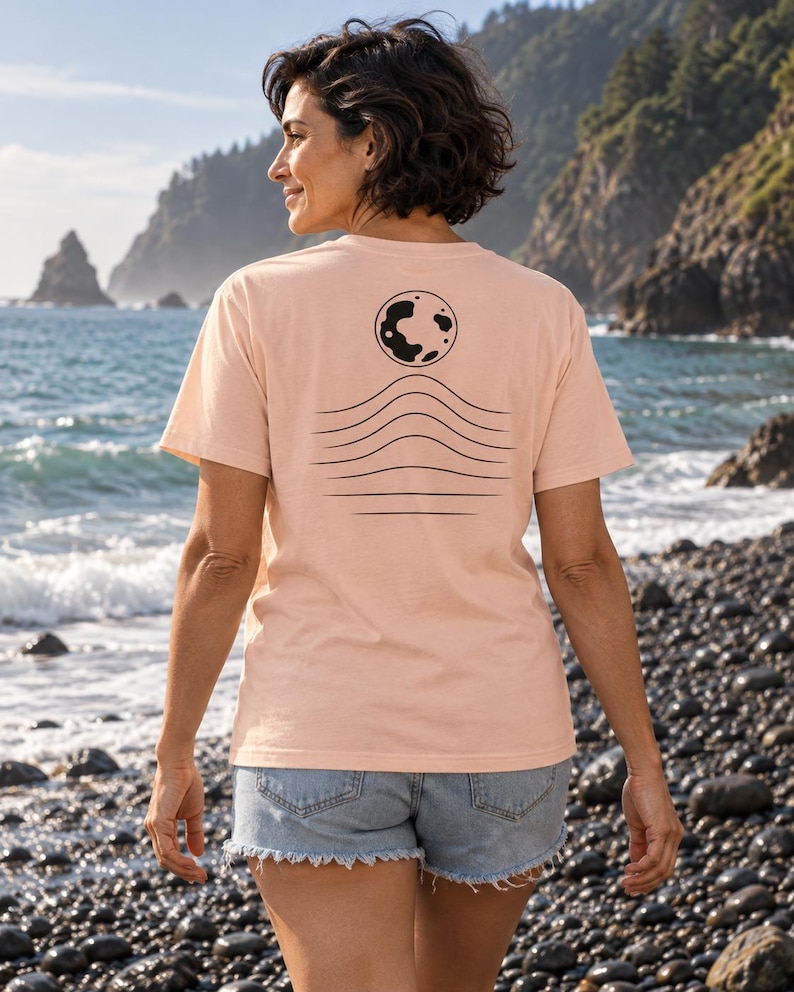 K&ouml;nnte beinhalten: Ein hellpfirsichfarbenes Kurzarm-T-Shirt mit einer schwarzen Grafik auf der R&uuml;ckseite. Das Design besteht aus einer kreisf&ouml;rmigen Mondform &uuml;ber einer Reihe von f&uuml;nf horizontalen, wellenf&ouml;rmigen Linien, die zum unteren Saum hin flacher werden.