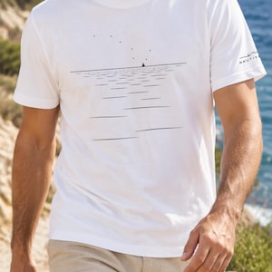 Peut inclure: Un t-shirt blanc &agrave; manches courtes orn&eacute; d'un graphisme au trait noir repr&eacute;sentant un horizon marin calme avec un petit voilier et des &eacute;toiles. La manche gauche porte l'inscription NAUTIY en noir avec une petite ic&ocirc;ne de vague au-dessus.