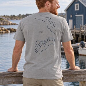 K&ouml;nnte beinhalten: Ein graumeliertes Kurzarm-T-Shirt mit einer dunkelblauen Linien-Grafik auf der R&uuml;ckseite. Das Design zeigt topografische H&ouml;henlinien und enth&auml;lt den Text -1200 m ATLANTIC, der in einer kleinen, klaren Schrift zwischen den Linien gedruckt ist.