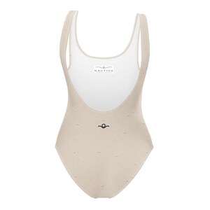 Peut inclure: Un maillot de bain une pi&egrave;ce beige avec un motif subtil de lignes ondul&eacute;es. Le v&ecirc;tement pr&eacute;sente une encolure d&eacute;gag&eacute;e et une doublure int&eacute;rieure blanche avec une &eacute;tiquette rectangulaire portant l'inscription NAUTIVE. Un petit logo noir est centr&eacute;.