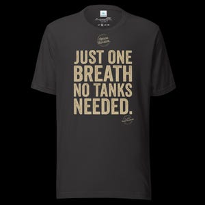 May include: A dark gray short-sleeve t-shirt featuring the beige text "JUST ONE BREATH NO TANKS NEEDED." with a circular "Apnea Division" logo above and a smaller "Est 2014 Pure Freedom" circular logo below the main text on the front.