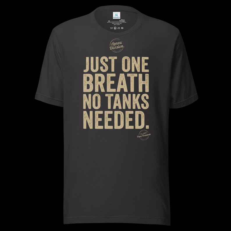 May include: A dark gray short-sleeve t-shirt featuring the beige text "JUST ONE BREATH NO TANKS NEEDED." with a circular "Apnea Division" logo above and a smaller "Est 2014 Pure Freedom" circular logo below the main text on the front.