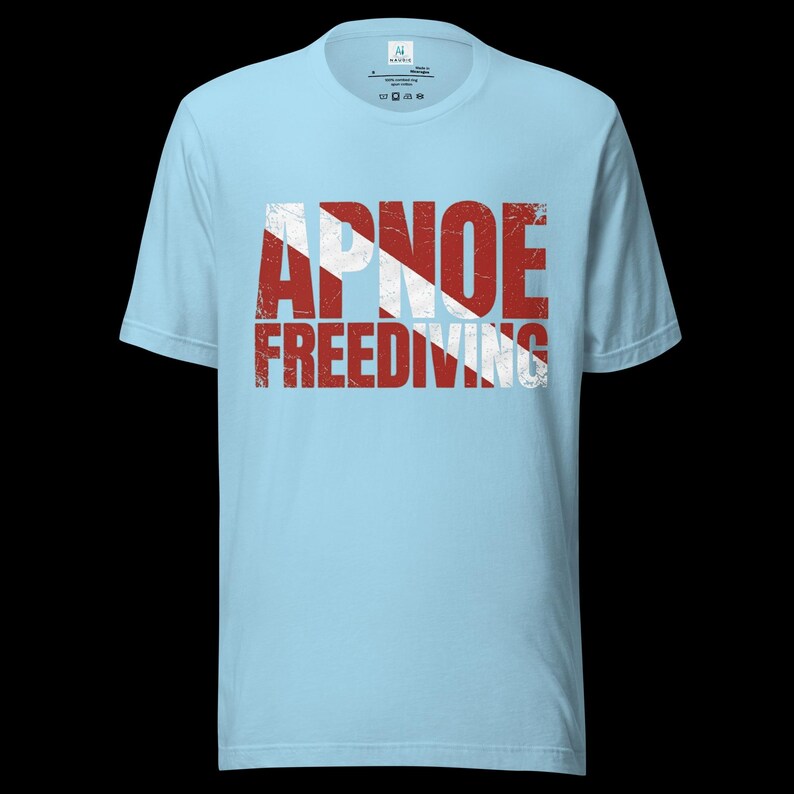 Puede incluir: Una camiseta de manga corta de color azul claro con el texto APNOE FREEDIVING en una fuente roja y blanca desgastada. El dise&ntilde;o incorpora una franja blanca diagonal sobre las letras rojas, imitando una bandera de buceo en el pecho.