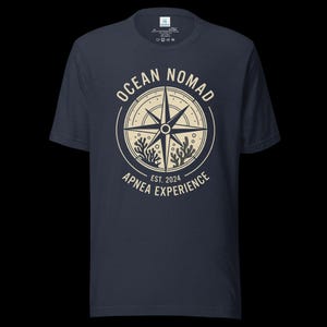 Op de afbeelding: Een donkerblauw t-shirt met korte mouwen en een cr&egrave;mekleurige cirkelvormige afbeelding op de borst. Het ontwerp bevat een kompasroos, koraalillustraties en de tekst "OCEAN NOMAD" en "APNEA EXPERIENCE EST. 2024" in een cirkelvormige lay-out.
