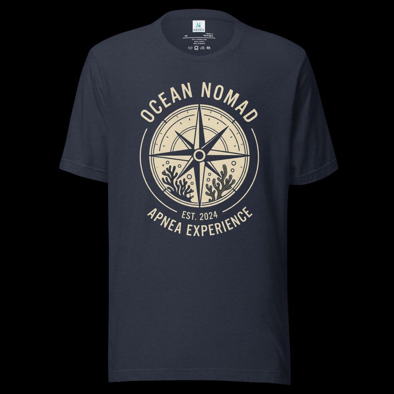Op de afbeelding: Een donkerblauw t-shirt met korte mouwen en een cr&egrave;mekleurige cirkelvormige afbeelding op de borst. Het ontwerp bevat een kompasroos, koraalillustraties en de tekst "OCEAN NOMAD" en "APNEA EXPERIENCE EST. 2024" in een cirkelvormige lay-out.