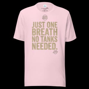 May include: A light pink short-sleeve t-shirt featuring the text "JUST ONE BREATH NO TANKS NEEDED." in a distressed tan font. Above the text is a circular "Apnea Division" logo, and below the text is a smaller circular "Est. 2019 Pure Freedom" logo.
