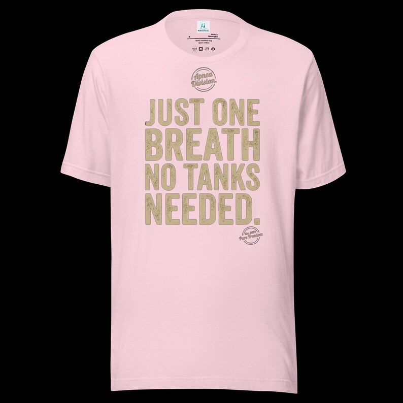 May include: A light pink short-sleeve t-shirt featuring the text "JUST ONE BREATH NO TANKS NEEDED." in a distressed tan font. Above the text is a circular "Apnea Division" logo, and below the text is a smaller circular "Est. 2019 Pure Freedom" logo.