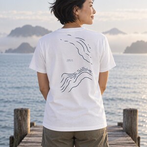 K&ouml;nnte beinhalten: Ein wei&szlig;es Kurzarm-T-Shirt mit einem dunkelblauen Grafikdruck auf der R&uuml;ckseite. Das Design besteht aus zwei S&auml;tzen wellenf&ouml;rmiger Linien, die die Ozeantopografie darstellen, mit dem Text -1200 m ATLANTIC in der Mitte dazwischen.