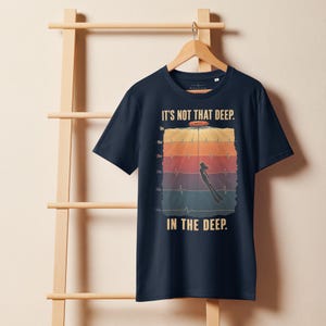 May include: A navy blue short-sleeve t-shirt featuring a graphic print of a diver descending through layered ocean depths. The design includes a heartbeat line, depth markers from 0m to 50m, and the text "IT'S NOT THAT DEEP. IN THE DEEP."