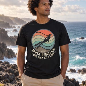 K&ouml;nnte beinhalten: Ein schwarzes Kurzarm-T-Shirt mit einer kreisf&ouml;rmigen Grafik in Retro-Sonnenuntergangsfarben wie Orange, Beige und Blaugr&uuml;n. Das Motiv zeigt einen Apnoetaucher sowie den Text "APNEA REBELLION", "EST. 2024" und "ONE KICK AT A TIME".