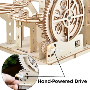 May include: A wooden marble run kit with gears, ramps, and a hand-powered drive mechanism. The kit is made of light-colored wood and features intricate cut-out designs. The text "Hand-Powered Drive" is visible on the image.