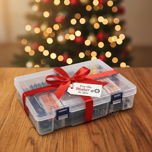 Könnte beinhalten: Eine transparente Plastikbox gefüllt mit elektronischen Bauteilen, gebunden mit einem roten Band und einem Anhänger mit der Aufschrift "For the Maker in You". Die Box steht auf einer Holzoberfläche, mit einem verschwommenen Weihnachtsbaum im Hintergrund.