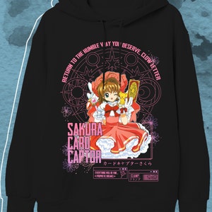 May include: Black hooded sweatshirt featuring a colorful graphic of a character from "Sakura Card Captor." The design includes the text "Sakura Card Captor" in pink and white, along with Japanese text and other phrases. The character is wearing a red dress.