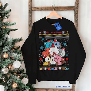 May include: Black crew neck sweatshirt with a colourful graphic of BT21 characters wearing Santa hats, and the text "BT21 CHRISTMAS". The sweatshirt is displayed on a wooden hanger.