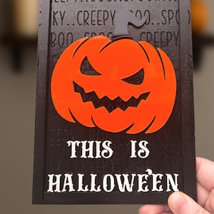 May include: A Halloween wooden sign featuring an orange pumpkin design and the words "THIS IS HALLOWEEN" in white. The black background has the words "creepy," "spooky," and "boo" etched into the wood, creating a spooky effect.