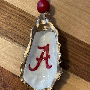 May include: Gold painted oyster shell ornament with a red "A" painted inside. The ornament has a red and gold bead at the top.