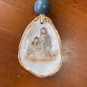 Può includere: Ornamento natalizio a conchiglia con una scena della Natività. Il guscio ha un bordo dorato ed è decorato con una perla blu e una perla dorata. La scena della Natività raffigura Maria, Giuseppe, Gesù bambino e un agnello.