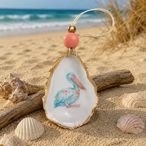 May include: An oyster shell ornament featuring a hand-painted blue pelican with pink floral accents. The shell has a gold-painted edge and is suspended by a jute twine loop adorned with a large peach-coloured wooden bead and a smaller gold bead.