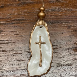 May include: A white oyster shell ornament with a gold cross design and gold beads hanging from a string.