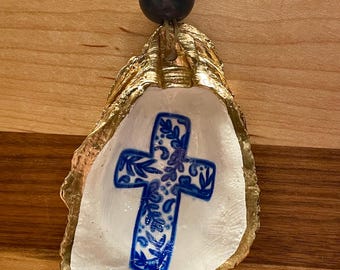 May include: A gold-rimmed oyster shell ornament with a blue and white cross design. The ornament is hanging from a blue bead and gold string.