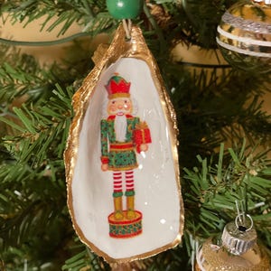 Puede incluir: Adorno navideño con una figura de cascanueces pintada dentro de una concha de ostra con bordes dorados. El cascanueces viste un uniforme rojo y verde y sostiene un regalo. El adorno cuelga de una cuenta verde y un hilo dorado.