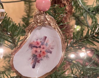 Puede incluir: Un adorno navideño de concha de ostra pintada de oro con fondo blanco y diseño de cruz floral. El adorno está colgado de un árbol de Navidad con cuentas rosas y doradas.