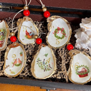 Op de afbeelding: Zes oester schelp ornamenten met gouden rand en geschilderde kersttaferelen. Elk ornament heeft een rode kraal en een nummer van 1 tot 6. De ornamenten worden gepresenteerd in een doos met bruine papieren strooisel.