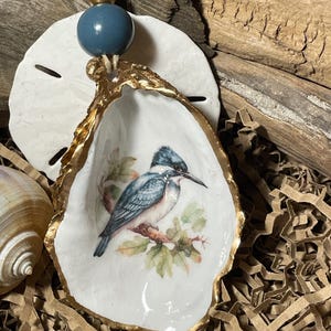 May include: An oyster shell ornament with a gold-painted edge, featuring a painted bird on a branch. The ornament is suspended by a string with a blue bead and a gold bead. The background includes a sand dollar and wood.