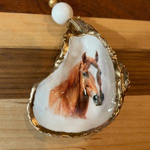 Könnte beinhalten: Ein dekoratives Austernschalen-Ornament mit einem gemalten Pferdeporträt. Die Schale hat einen goldfarbenen Rand und ein weißes Innere. Eine weiße Perle und eine goldfarbene Perle sind oben mit einer Schnur befestigt.