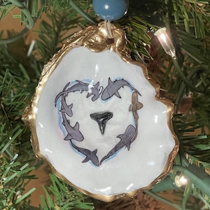May include: A unique Christmas ornament made from a white oyster shell with a gold-painted edge. The shell features a heart-shaped design of gray sharks and a black shark tooth in the center. A blue bead and gold bead are attached for hanging.