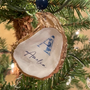 May include: A gold-painted oyster shell ornament with a blue and white floral letter "A" and the name "Amelia" written in cursive. The ornament is hanging on a Christmas tree.