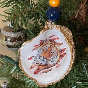 May include: A hand-painted oyster shell Christmas ornament featuring a tiger's face. The shell has a gold-painted edge and is suspended by a string with blue and orange beads. The ornament hangs on a Christmas tree.