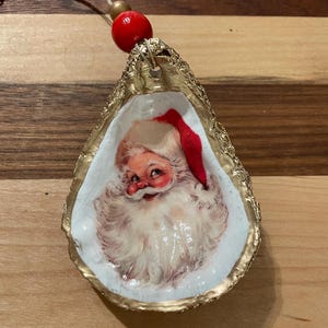 Könnte beinhalten: Ein goldbemalter Austernmuschel-Weihnachtsanhänger mit einem Vintage-Bild von Weihnachtsmann im Inneren. Der Anhänger hat eine rote Perle oben zum Aufhängen.