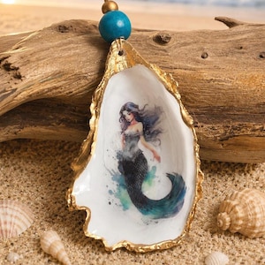 May include: A decorative oyster shell ornament featuring a painted mermaid with flowing dark hair and a teal tail. The shell edges are gilded in gold, and it hangs from a jute string adorned with a single blue wooden bead and a smaller gold bead.