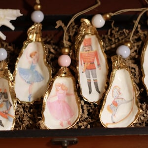 Puede incluir: Adornos de concha de ostra con bordes dorados, cada uno con ilustraciones temáticas de ballet. Los adornos están adornados con cuentas y cuerda para colgar, y se exhiben en una caja de madera.