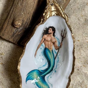 May include: A decorative oyster shell ornament featuring a hand-painted merman with long dark hair holding a trident. The shell has a gold-painted edge and is topped with a blue bead and a small gold bead on a natural fiber cord loop.