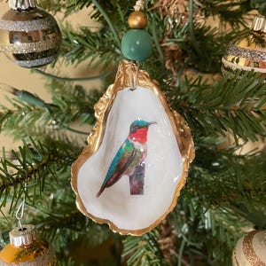 May include: An oyster shell Christmas tree ornament with a painted hummingbird design. The shell has a gold-painted edge and is hung by a twine string with green and gold beads. Other baubles are visible on the tree.