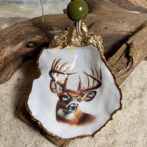 May include: A decorative oyster shell ornament featuring a detailed image of a brown deer with antlers. The shell has a gold-painted scalloped edge and is suspended by a jute twine string adorned with a large olive green bead and a smaller gold bead.