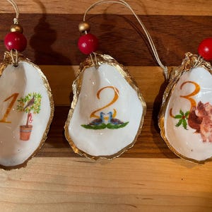 Op de afbeelding: Drie oesterschelpversieringen met gouden randen, elk met een nummer en een illustratie. De nummers zijn goudkleurig en de illustraties omvatten een potplant, twee vogels en drie eekhoorns. Elke versiering heeft een rode kraal.