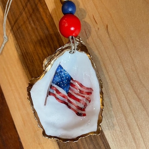 Puede incluir: Adorno de concha de ostra con una bandera americana pintada. La concha tiene un borde dorado y está ensartada con un cordón natural y tres cuentas de madera en rojo, azul y dorado. Una decoración patriótica.