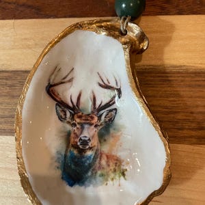 May include: An oyster shell trinket dish with a gold-painted rim. The interior features a watercolor-style image of a deer with antlers. A green bead and a gold bead are attached to the shell with a string.