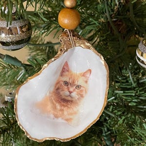 May include: A handmade Christmas ornament featuring a ginger cat portrait inside a white oyster shell with a gold-painted edge. The ornament is suspended by a string and wooden beads, ready to hang on a Christmas tree.