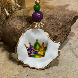 May include: A white oyster shell ornament with a gold-painted edge, featuring a colorful crown design. The ornament is adorned with a string of wooden beads in gold, green, and purple. A unique, handcrafted decorative item.