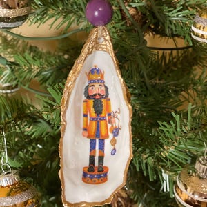 Peut inclure: Ornement de Noël représentant un casse-noisette peint à la main à l'intérieur d'une coquille d'huître dorée. Le casse-noisette est habillé en bleu et jaune, avec une perle violette et une perle dorée en haut pour l'accrocher. L'ornement est sur un sapin de Noël.