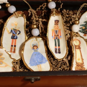 Puede incluir: Adornos decorativos de concha de ostra con borde dorado, cada uno con una ilustración diferente con el tema Cascanueces. Los adornos están ensartados con hilo y cuentas, y se presentan en una caja de madera con relleno de papel.